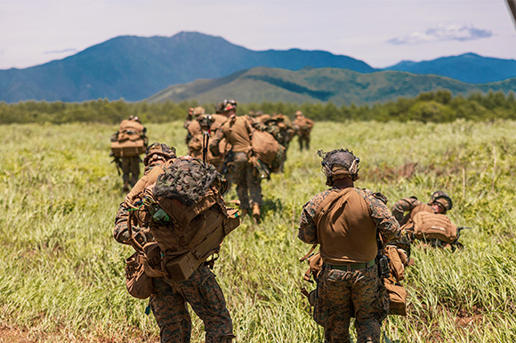 Marines trekking through tall grass with lush mountains in the distance.