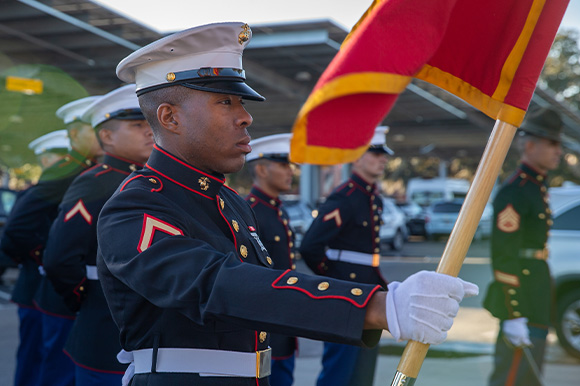 A Marine in dress uniform with his arm outstretched, holding the USMC flag.