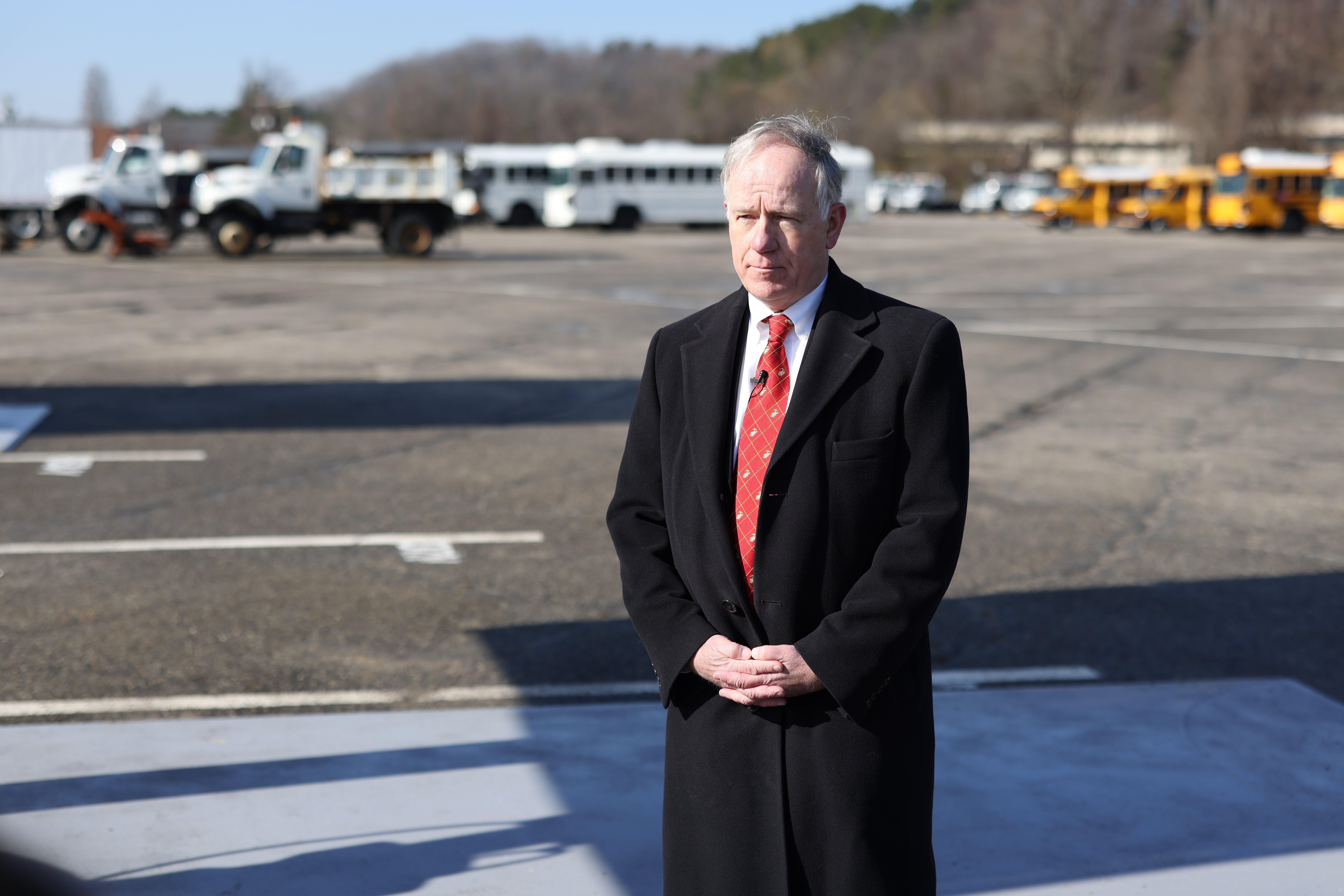 A solemn man in a black overcoat, standing in a parking lot with his hands folded.