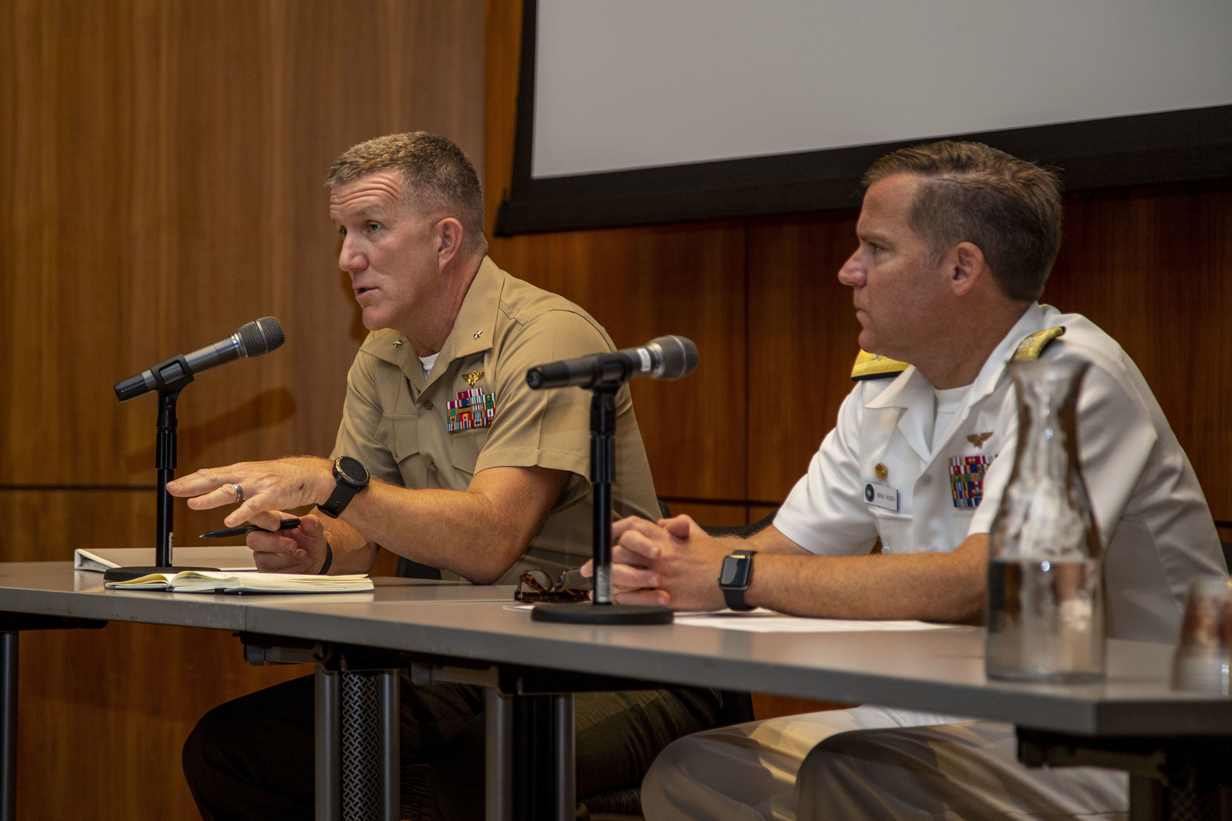Two men sitting at a table on stage, speaking into microphones to, presumably, an audience.